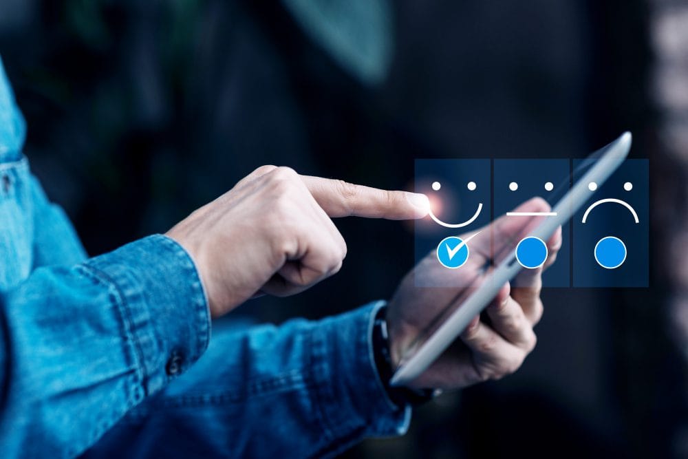 Una persona utiliza una tableta y selecciona un icono de cara feliz con una marca de verificación en un panel de opciones de respuesta -impulsado por IA en la experiencia del cliente para pymes- que también incluye iconos de caras neutras y tristes.